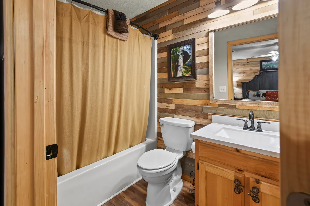 A warmly-lit bathroom with a wooden accent wall, a vanity sink, framed artwork, and a soft shower curtain. The space exudes relaxation and offers all the comforts needed for self-care routines.