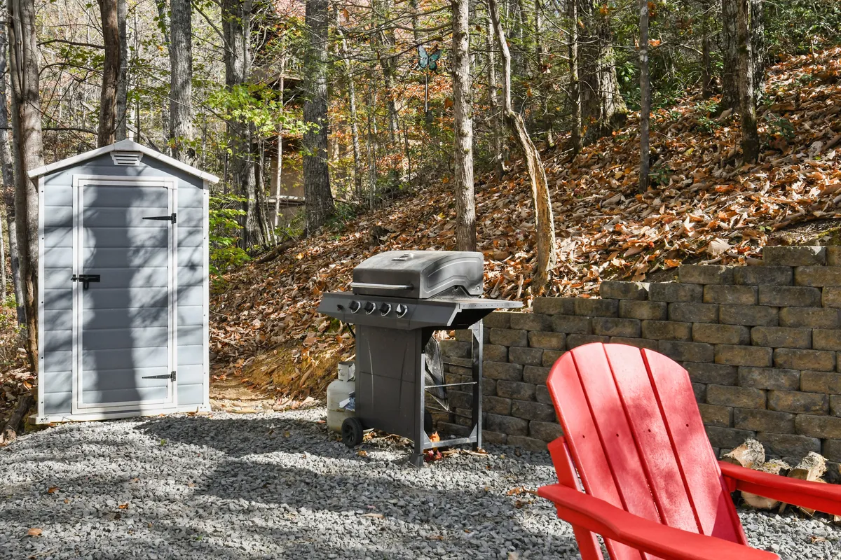 This image features a gray storage shed next to a grill and a red Adirondack chair, all set against a stone wall in a backyard. The outdoor space suggests a relaxed environment for barbecuing, hanging out, or enjoying the fresh mountain air with friends or family.