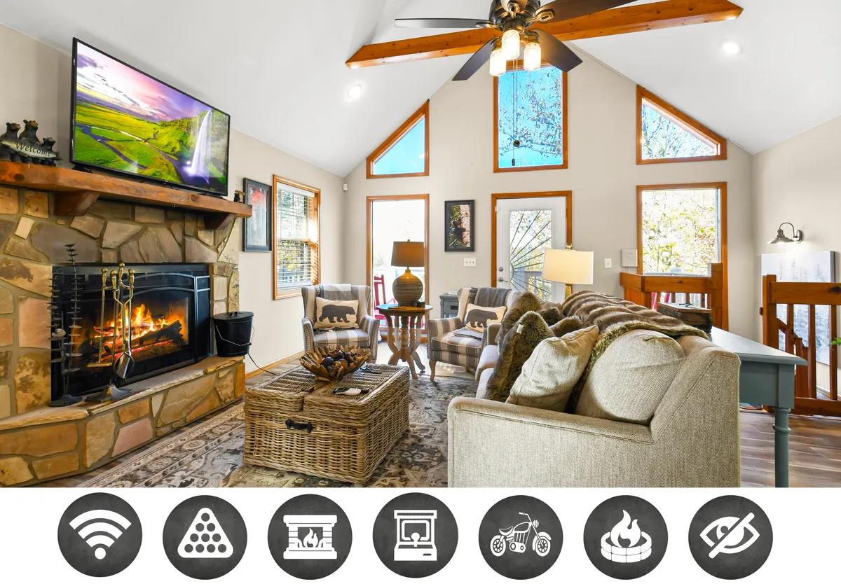 The image showcases a cozy cabin living room, featuring a glowing wood-burning fireplace as the centerpiece, with comfortable seating arranged around a rustic coffee table. Large windows offer glimpses of the natural surroundings, filling the space with warm, inviting light. A TV is mounted nearby, and the ambiance is perfect for relaxing, sharing stories, or enjoying a game night.