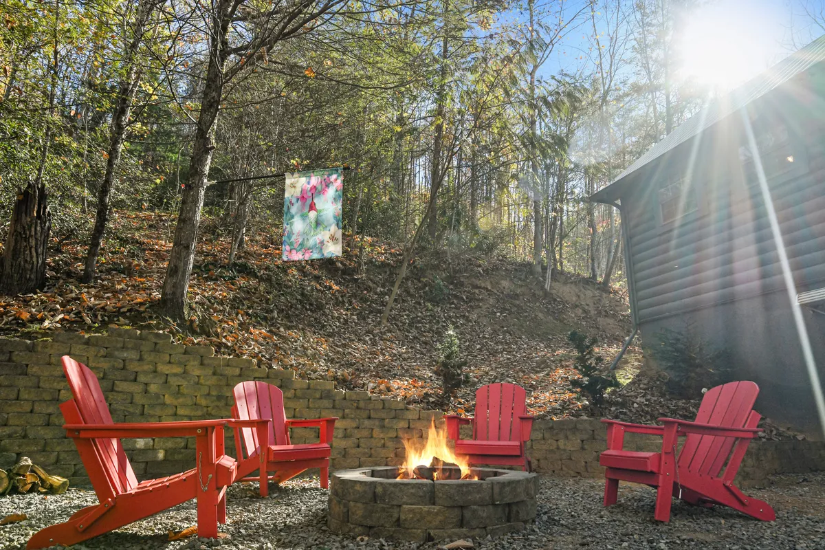 An inviting outdoor scene featuring a fire pit surrounded by red Adirondack chairs, with a colorful fabric banner adding a playful touch to the natural setting. The area is nestled amid lush greenery, creating an ideal spot for evening gatherings, marshmallow roasting, or storytelling under the stars.