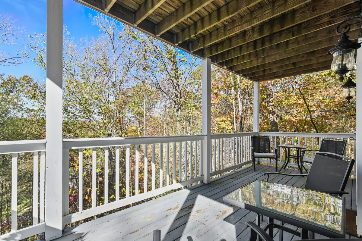 A tranquil exterior dining space is featured here, with a sturdy glass table and black chairs set atop a wooden deck surrounded by a natural woodland backdrop. The wooden railing frames the space, and the table offers a perfect spot for al fresco meals. The atmosphere is peaceful and integrates seamlessly with the forest surroundings, inviting group gatherings or quiet contemplation.