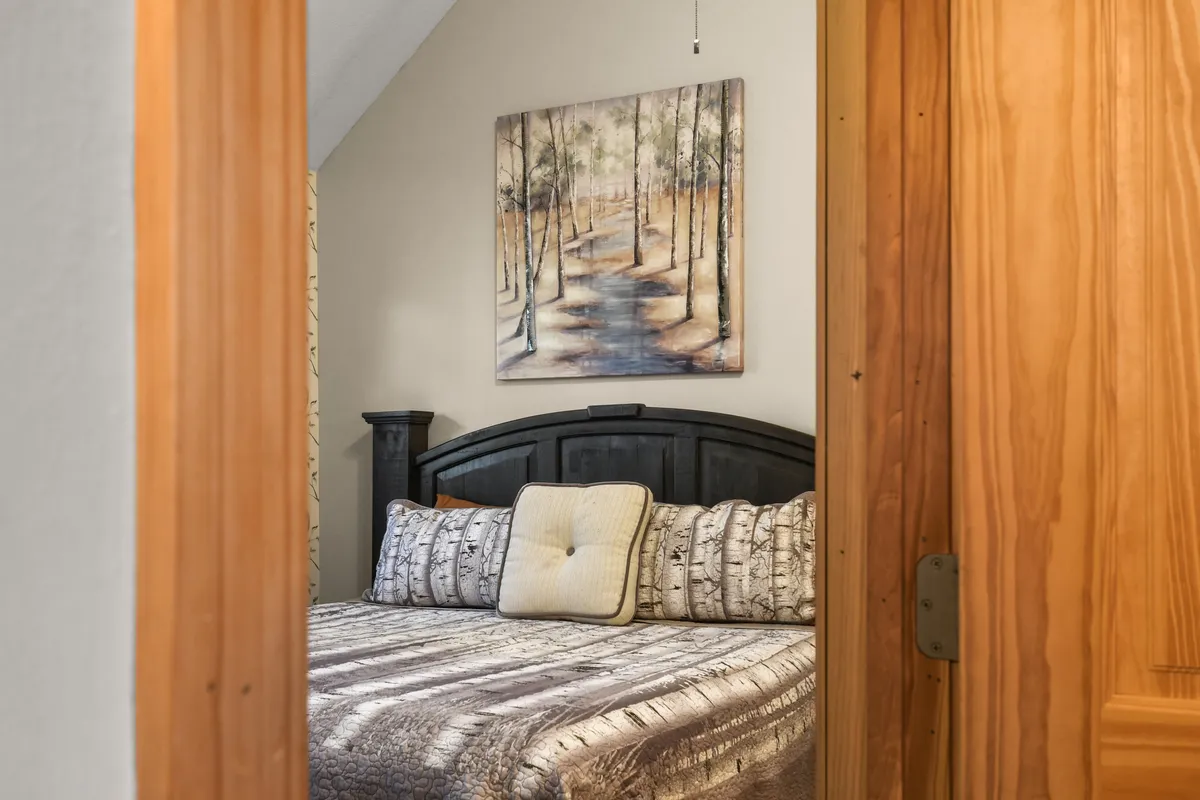 A tranquil bedroom scene with a bed adorned in decorative pillows, a wooden headboard, and soothing artwork of a forest path. The atmosphere feels serene and restful, ideal for unwinding.