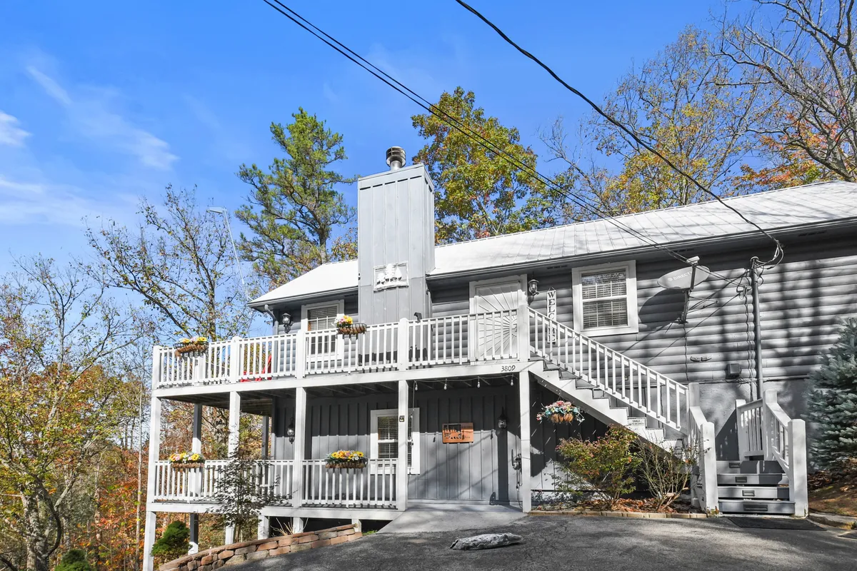 A bright and welcoming front porch with steps leading up, planters filled with flowers, and ample space for group gatherings. The setting is perfect for hosting seasonal get-togethers or taking in the scenic surroundings at the cabin's entrance.