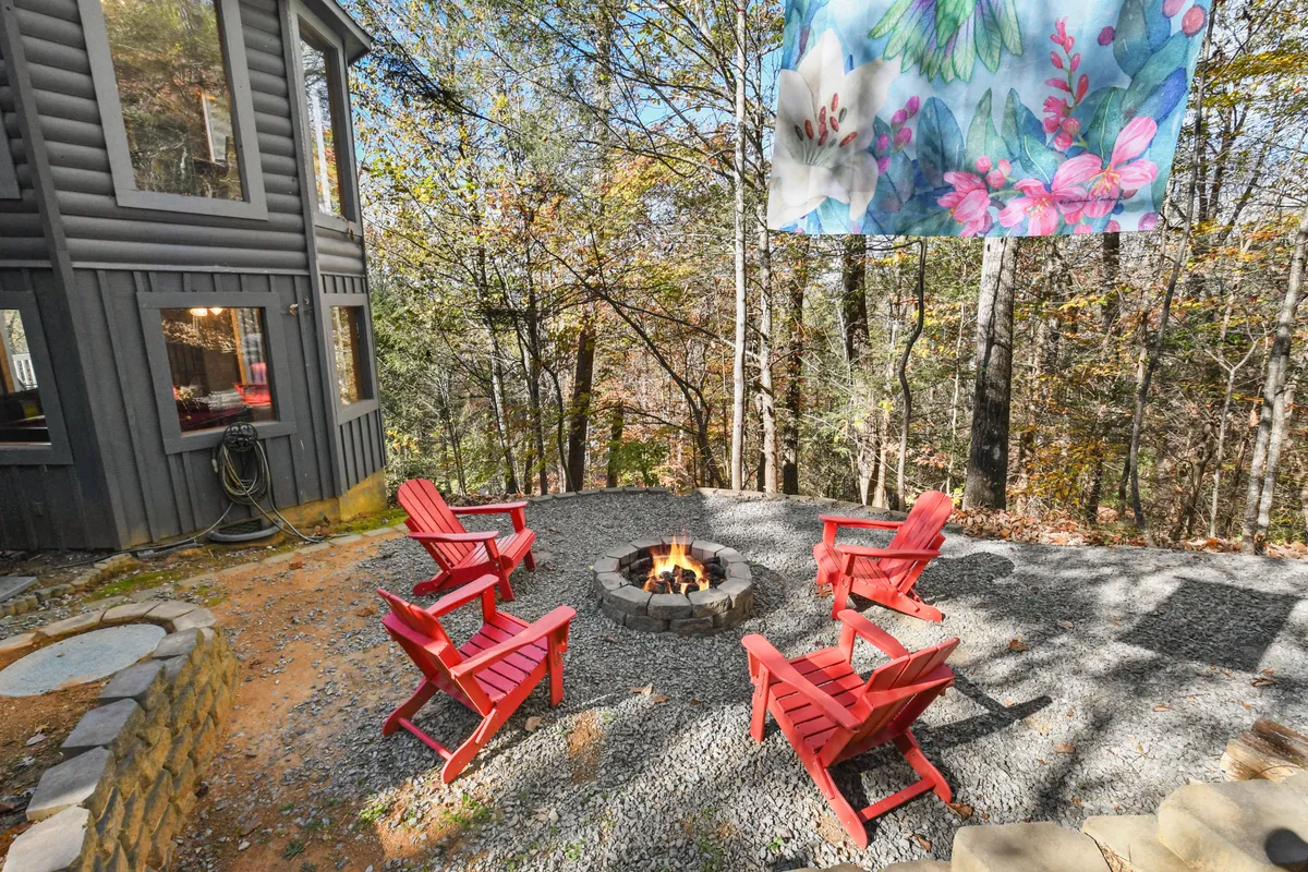 A cozy fire pit scene with a stone ring surrounded by red chairs, set in a natural outdoor clearing. A decorative banner adds a festive touch, while the setting sun bathes the area in a warm glow. The arrangement suggests evenings filled with stories, laughter, and roasted marshmallows, capturing the spirit of communal outdoor relaxation.