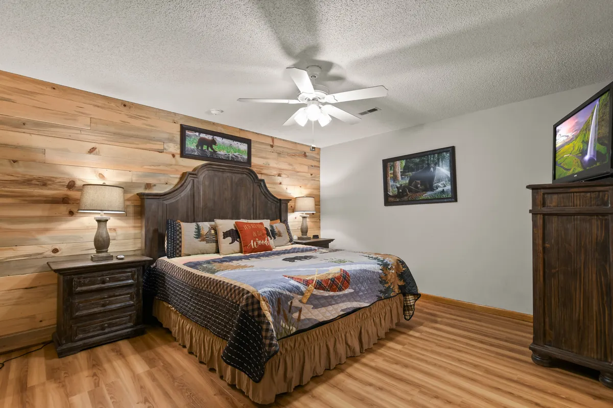 A cozy bedroom featuring a wooden headboard, colorful quilt, a nightstand with a lamp, and wall art. Natural light filters into the space, highlighting the restful ambiance.
