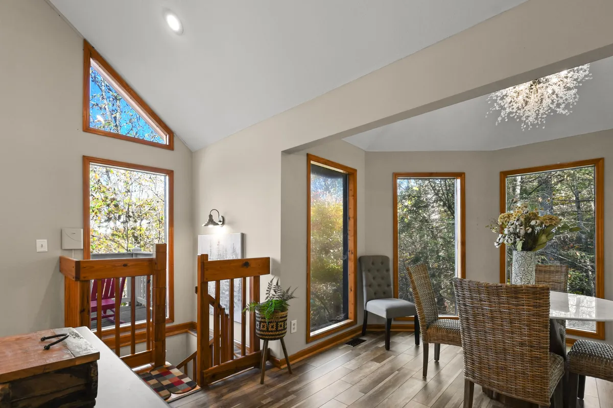 A bright dining area with a floral centerpiece on the table, large windows letting in ample natural light, and a wooden staircase leading to an upper level. The arrangement suggests a space perfect for casual dining, morning coffee, or group celebrations.