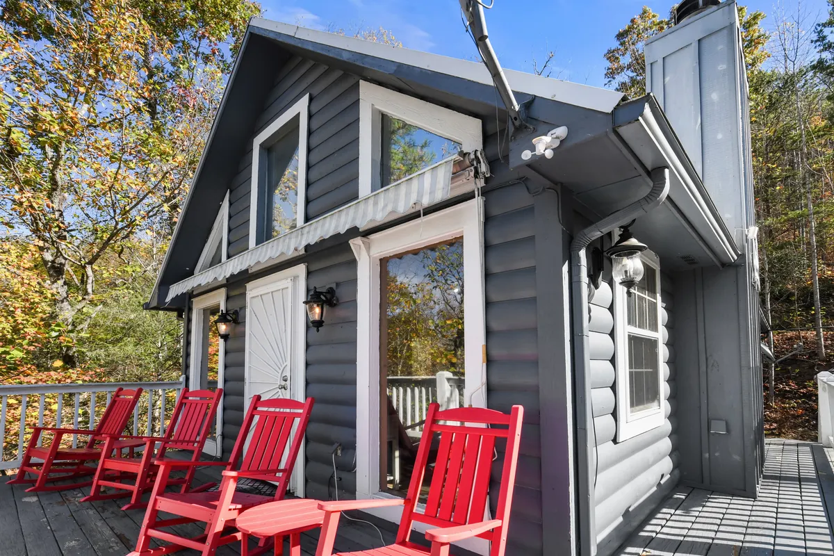 This image displays a cozy deck area with two inviting red rocking chairs facing outward, perched in front of a gray log cabin exterior with large windows. The deck overlooks lush greenery, suggesting a tranquil tree-lined setting. The scene radiates warmth and relaxation, making it easy to imagine enjoying a sunrise coffee or an evening chat under the stars.