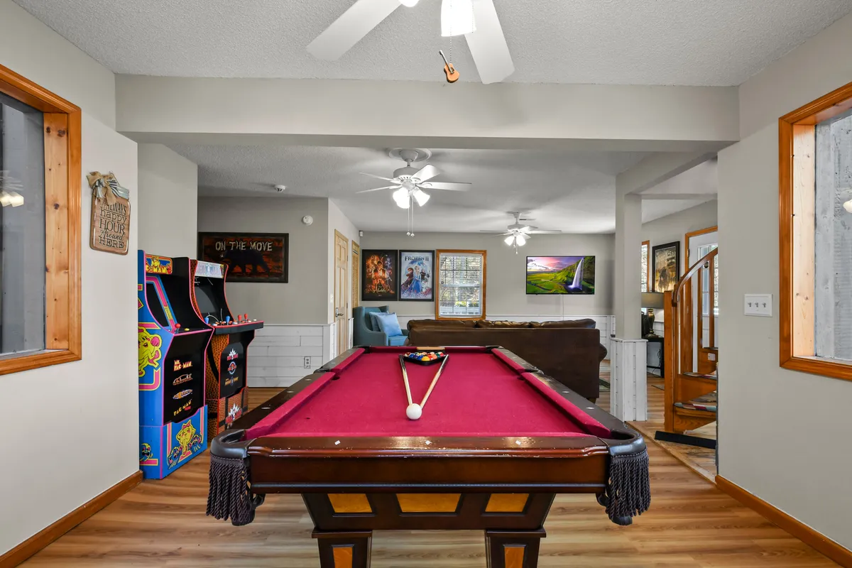 This image captures the central area of the game room, featuring a pool table, an arcade machine, and a comfortable seating area. The space is accented with playful artwork on the walls, and the arrangement invites both active play and relaxed socializing. Natural light streams in, giving the room an open and lively feel.
