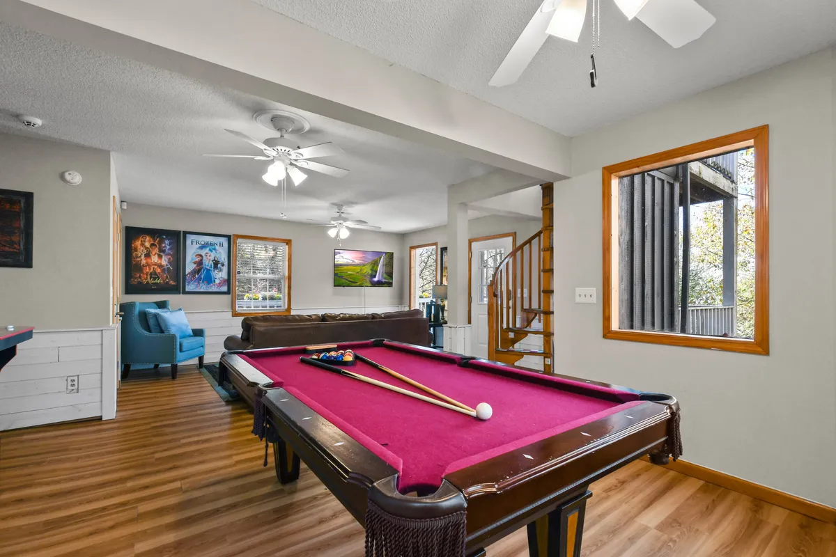 A view of the game room from a different angle, showcasing the pool table, a spiral staircase, and a plush seating area for watching movies or relaxing after gameplay. The overall mood is casual and welcoming, ideal for gatherings and family movie nights.