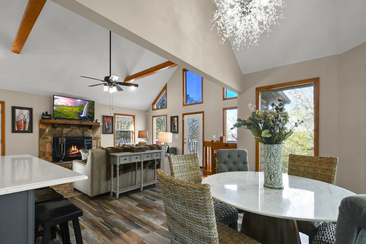A warm, open-concept living and dining space, featuring a wood-burning fireplace as the focal point. Large windows bathe the area in natural light, while a spacious dining table sits nearby, perfect for shared meals. Artworks on the wall add a touch of character and homeliness.