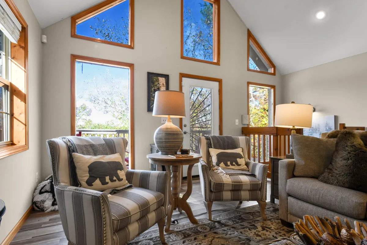 A cozy corner with striped armchairs featuring bear motifs, a decorative lamp, wooden table, and large windows granting a scenic view. The space exudes warmth and invites intimate conversation or peaceful moments shared over coffee.